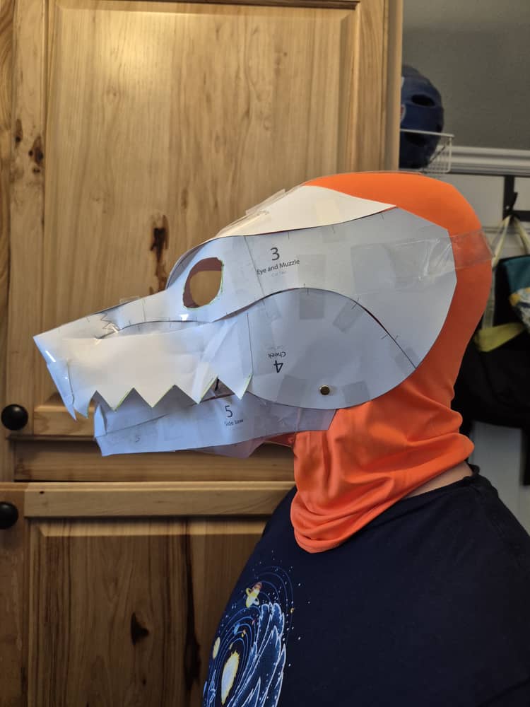 A side view of a man wearing an orange hood with a white paper mask with a long snout and jagged teeth.
