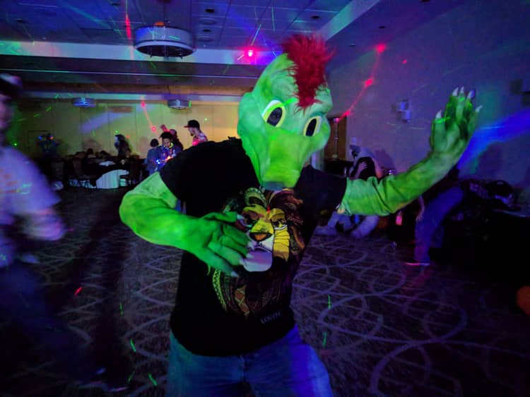Ine's fursuit, a green lizard with red feathers on his head, dancing in a dimly lit ballroom with colorful lights with other people dancing in the background.