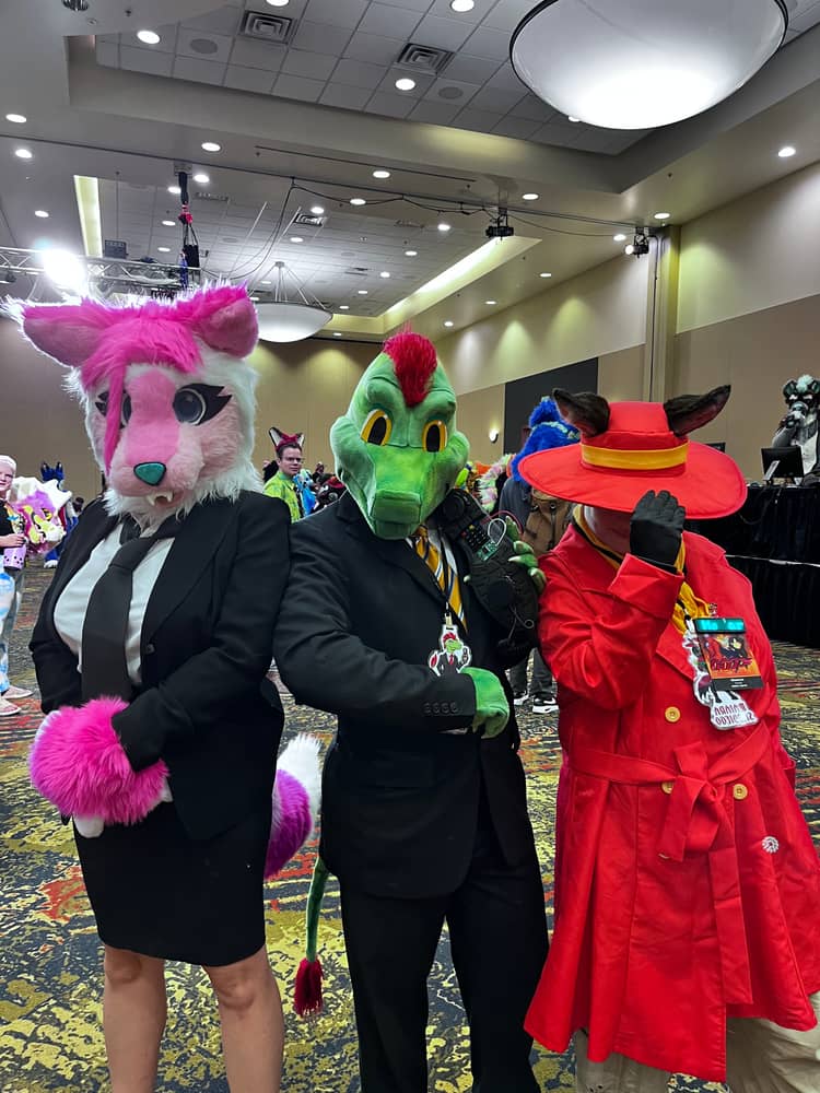 Ine's fursuit, a green lizard with red feathers on his head with a pink feline, both wearing black suits, and a woman in a bright red Carmen Sandiego trenchcoat and hat with horse ears.