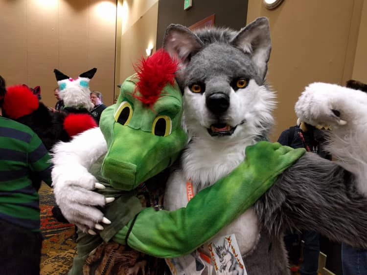 Ine's fursuit, a green lizard with red feathers on his head, hugging a gray wolf fursuiter who is flexing with one arm.