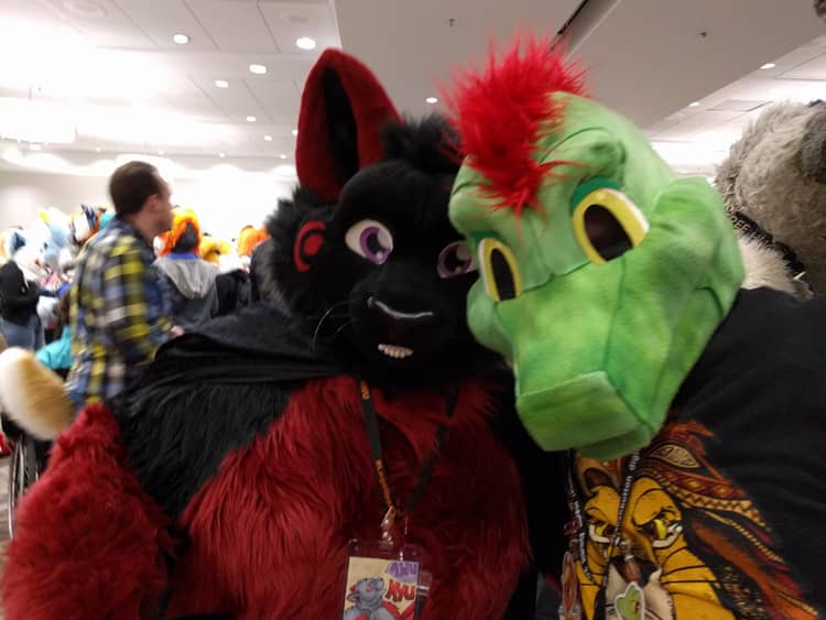 Ine's fursuit, a green lizard with red feathers on his head, standing next to a fursuit of a black cat with red markings on her ears, paws, tummy, and cheek.